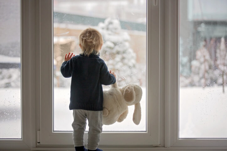 A small boy with a large stuffed rabbit dangling from one hand, the other hand pressed against the patio door, watching the yard fill up with snow.