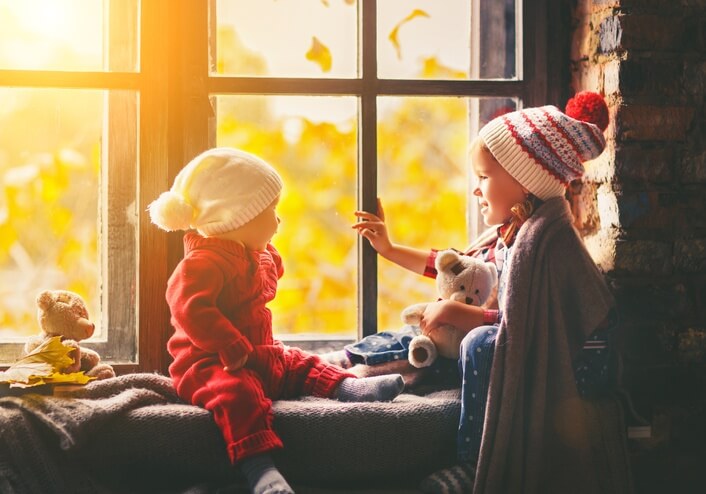 Two small children wrapped in cozy blankets and winter hats sitting in the window seat, holding stuffed animals.