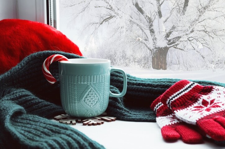 A cup of hot cocoa with a candy cane sitting on the windowsill next to a pair of gloves and a scarf, as the snow falls outside the window.