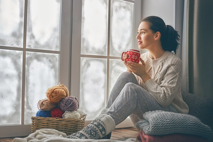 A woman sitting in the window seat with a hot cup of tea next to a stack of neatly folded hand-knit blankets and a basket filled with balls of yarn.