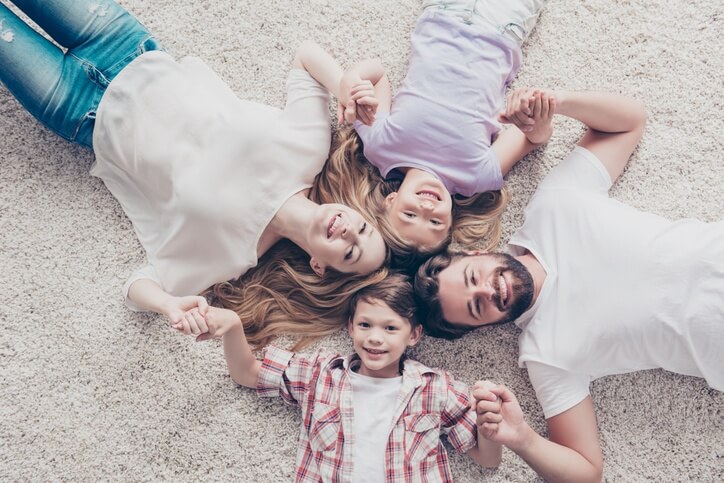 A smiling family of 4 relaxes on the floor of their air-conditioned home in a pinwheel formation, head-to-head, holding hands.